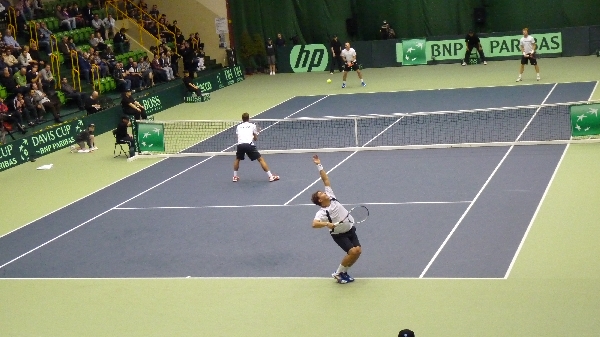 Deviscup - hala główna przekształcona w kort tenisowy. Mecz debla mężczyzn. Na pierwszym planie zawodnik serwujący piłkę.